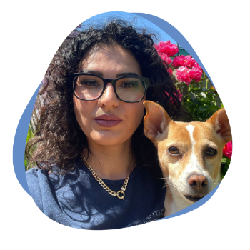 Junge Frau mit dunklen lockigen Haaren, einer Brille und einem Hund auf dem Arm